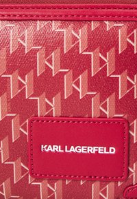 Crvena teksturirana torba s geometrijskim uzorkom i glatkom kožnom oznakom s utisnutim natpisom "KARL LAGERFELD" u bijeloj boji.