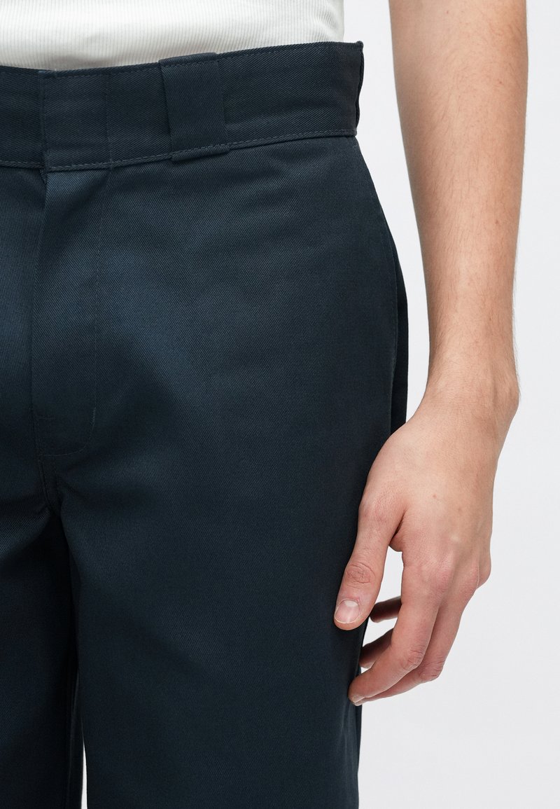 Dunkelblaue Hose mit glatter Textur, geradem Schnitt und flachem Bund. Verfügt über minimale Nähte und eine Hand, die an der Seite ruht.