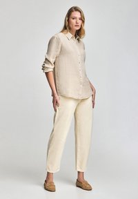 Camisa de lino beige con cuello y cierre de botones, combinada con pantalones color crema y zapatos de tela marrón claro. Ajuste simple y relajado.