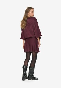 Femme aux longs cheveux portant une robe bordeaux, des collants noirs et des bottes noires à boucle mi-mollet, se tenant debout et regardant par-dessus son épaule.