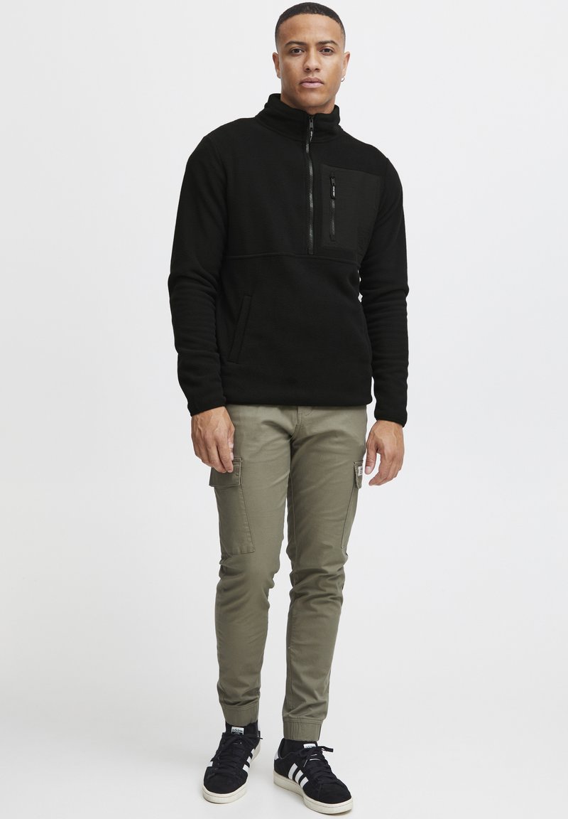 Svart fleece genser med høy krage, halv glidelås og frontlomme, kombinert med olivengrønne cargo-bukser og svarte sneakers.