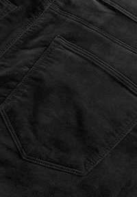 Black corduroy fabric, featuring a textured surface, with a visible back pocket in a standard diamond shape and stitched detailing.