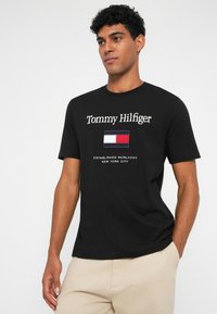 Mustang mustari T-särk, millel on "Tommy Hilfiger" tekst ja ristkülikukujuline logo punase, valge ja sinise osaga, ümar kaelus, lühikesed varrukad.