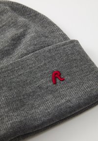 Bonnet tricoté gris avec un revers, arborant une petite lettre "R" brodée en rouge sur le côté. Texture douce, design simple.