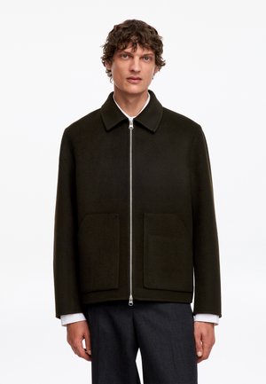 Homme portant une veste zippée vert foncé avec de grandes poches avant, poignets de chemise blancs visibles et pantalon gris foncé, debout devant un fond uni.