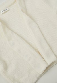 Hvid cardigan i blødt, struktureret stof med V-hals og knapdetaljer, der har et subtilt vaffelmønster. Mærket er synligt indeni.