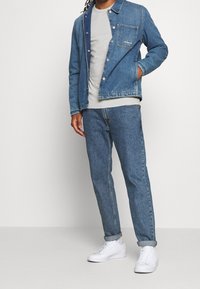 Muž v modré džínové bundě a džínách, světle šedém tričku a bílých teniskách, stojí s jednou rukou v kapse bundy.