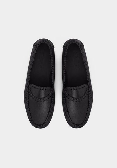 Ζευγάρι μαύρων δερμάτινων μοκασίνων χωρίς κορδόνια με πλεκτή διακόσμηση γύρω από τις άκρες, εμφανιζόμενα από πάνω σε απλό φωτεινό φόντο.
