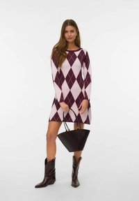 Donna che indossa un vestito argyle viola e rosa chiaro, tiene una borsa nera e indossa stivali da cowboy marrone scuro, in piedi su uno sfondo bianco.
