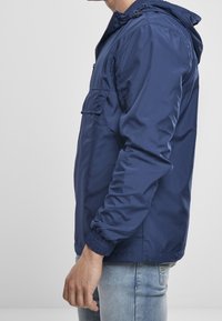 Granatowa, lekka kurtka z kapturem, elastycznymi mankietami, kieszenią z przodu i gładką teksturą, noszona z jasnymi dżinsami.