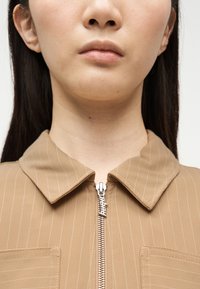 Κοντινό πλάνο ενός ατόμου που φοράει μπεζ πουκάμισο με ρίγες και φερμουάρ, με γιακά, δείχνοντας το κάτω μέρος του προσώπου και τον λαιμό.