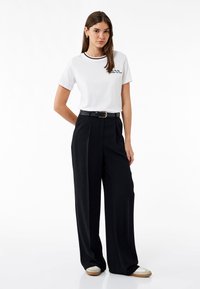 Camiseta blanca con bordado negro y cuello en contraste, combinada con pantalones negros de talle alto, con pierna ancha y cinturón negro.