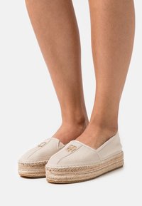 Baskets espadrilles beige avec semelles en jute tissé, tige en tissu texturé et un petit logo brodé à l'avant. Design plat, à enfiler.