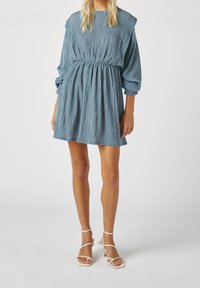 Vestido azul texturizado con un corte holgado, cintura fruncida y mangas largas. Combinado con sandalias de tacón blanco con tiras.