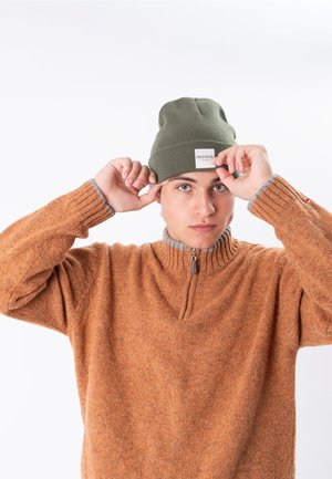 Young person adjusting green beanie with "OCCULTO" label, wearing orange zip-up sweater against plain white background.