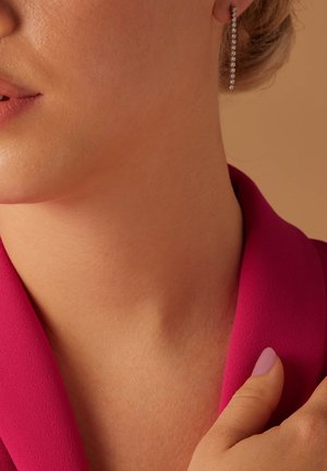 De longues boucles d'oreilles fines en argent avec de petites pierres précieuses transparentes, accompagnées d'un col en tissu rose. Le teint est naturel et lisse.
