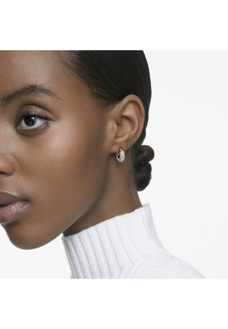 Zlaté kruhové náušnice s textúrovaným povrchom a zakomponovanými trblietavými kamienkami. Modelka má hladký účes a nosí biely rolák s rebrovanou štruktúrou.