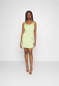 Vestido mini em satin verde claro com alças finas e cintura ajustada. Apresenta um bolso lateral e uma textura suave, complementado por saltos transparentes.