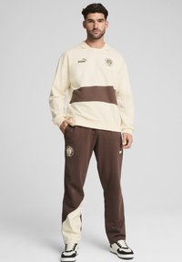 Cremefarbener Sweatshirt mit einem braunen horizontalen Streifen, kombiniert mit braunen Hosen, die ein kontrastierendes cremefarbenes Panel aufweisen. Gesticktes Logo und Branding.