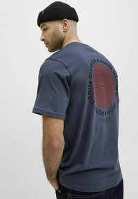 Krátký rukáv námořnické tričko vyrobené z bavlny, s kulatou červenou grafikou a černým textem na zadní straně. Model také nosí černou čepici.