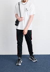 Vit t-shirt med svarta grafiska tryck, svarta träningsbyxor med röda sidostripes och svarta sneakers med grå detaljer. Liten svart axelremsväska.