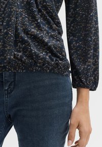 Langärmlige Bluse aus dunkelblauem Stoff mit einem subtilen beigen und weißen abstrakten Muster; geraffter Taillenbund und elastische Bündchen; kombiniert mit dunklen Jeans.