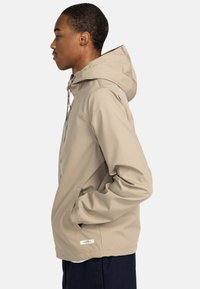 Beige Kapuzenjacke mit glatter Textur, ausgestattet mit Seitentaschen und verstellbaren Kordelzügen. Einfache Gestaltung mit lässiger Passform.