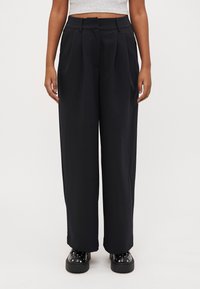 Wide-leg black trousers with pleats, made from a textured fabric. Features belt loops and a relaxed fit, paired with chunky black shoes.