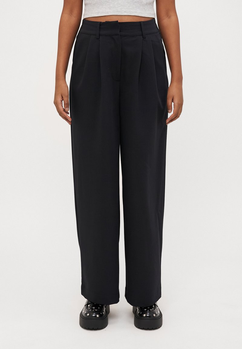 Wide-leg black trousers with pleats, made from a textured fabric. Features belt loops and a relaxed fit, paired with chunky black shoes.