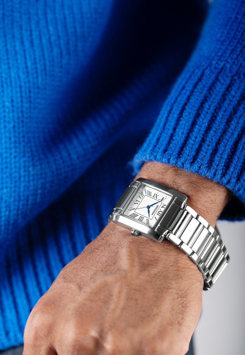 Montre carrée en argent avec un cadran blanc, des accents bleus et des chiffres romains, assortie d'un bracelet en métal argenté. Portée sur une main contre un pull bleu.