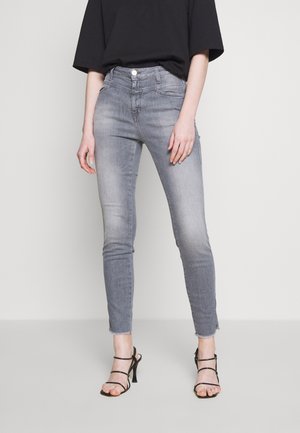 Mujer con jeans pitillo gris descoloridos con dobladillo sin rematar y sandalias negras con tiras y tacón, de pie contra un fondo liso de color claro.