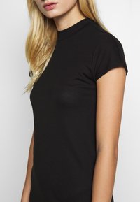 Blondynka ubrana w dopasowany czarny t-shirt z krótkim rękawem i wysokim dekoltem, przedstawiona od połowy torsu do ust na białym tle.