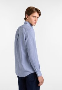 Ung man med ljust brunt hår iklädd en blå och vit rutiga långärmad skjorta och mörkblå byxor, som tittar över sin axel.