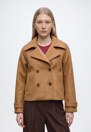 Jeune femme aux longs cheveux blonds portant un manteau croisé beige sur un pull rouge et un pantalon marron foncé, debout devant un fond clair.