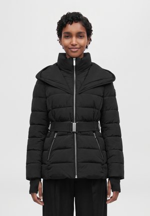Une femme porte une doudoune noire matelassée à col montant, avec une fermeture éclair sur le devant, une ceinture et des poches zippées, sur un fond uni.
