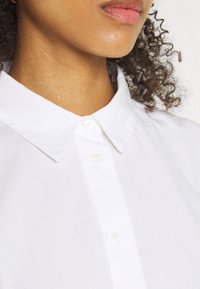 Camicia bianca con colletto, caratterizzata da una chiusura a bottoni, realizzata in tessuto leggero con una texture liscia e un design classico.