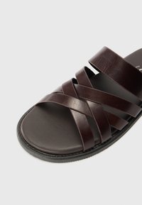 Sandale en cuir marron avec des brides entrecroisées, finition lisse et semelle plate. Design minimaliste sans accents supplémentaires.