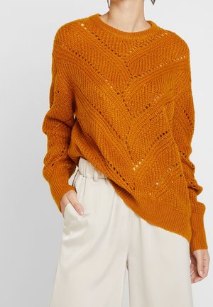 Rust-oranje grofgebreide trui met chevronpatroon, gedragen met crèmekleurige broek met hoge taille, één hand in de zak, op een neutrale achtergrond.