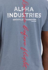 Camiseta de algodón gris con un atrevido texto blanco "ALPHA INDUSTRIES", y con letras rojas "Retirar antes del vuelo" en vertical a lo largo del costado.