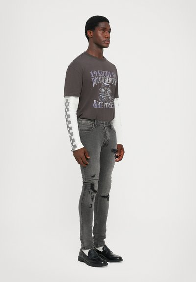 Graues Grafik-T-Shirt mit kurzen Ärmeln, das einen Druck und ein weißes Langarmunterhemd zeigt. Ausgewaschene graue Skinny-Jeans mit Distressed-Details und schwarzen Schuhen.