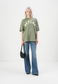 Overgrote olijfgroene t-shirt met witte "REPLAY" tekst, gecombineerd met uitlopende blauwe jeans en een zwarte geperforeerde handtas.