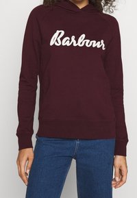 Bordový mikina s bíle vyšitým logem "Barbour". Obsahuje klokankovou kapsu, žebrované manžety a volný střih. Materiál se zdá být měkký.