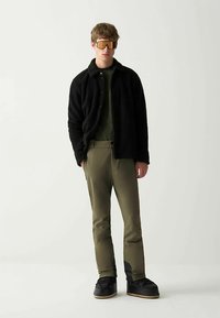 Veste en polaire noire, chemise verte, pantalon olive avec accents aux genoux, et chaussures noires. De grandes lunettes de soleil teintées dorées complètent le look.