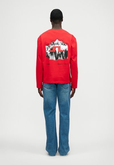 Red long-sleeve shirt featuring a mountain graphic and logo on the back, paired with blue flared jeans. Simple design, casual fit.