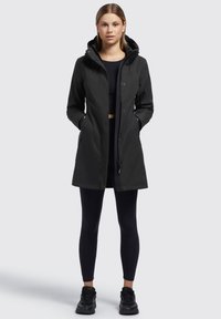 Parka imperméable noir avec capuche, dotée de boutons à l'avant, de poches et d'une coupe droite. Portée sur des leggings noirs et des baskets.