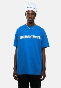 Camiseta de algodón azul con "GRIMEY BOYS" en letras blancas y en negrita, combinada con un gorro blanco que presenta el mismo logo, llevada con pantalones negros.