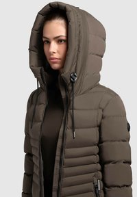 Chaqueta marrón acolchada con una gran capucha, diseño acolchado vertical, ajustes con cordón y bolsillos laterales con cremallera. Textura suave.