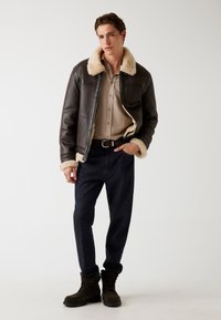 Giacca in pelle marrone con colletto e polsini in pelliccia beige, camicia beige con bottoni, pantaloni in denim scuro e stivali marroni con lacci.