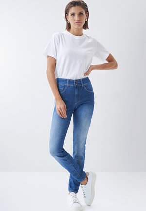 Jeans de tiro alto azul con un ligero desvanecimiento, detalles de costura en los bolsillos, combinados con una camiseta blanca lisa y zapatillas blancas.