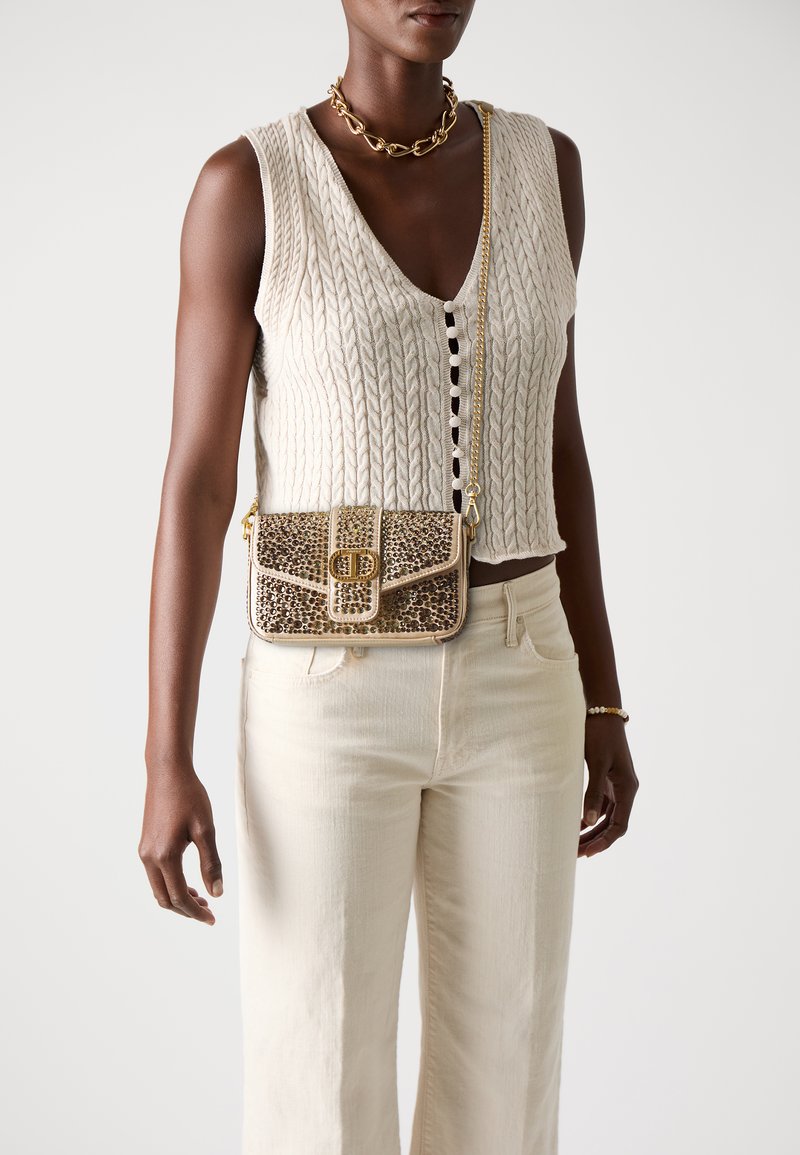 Gilet en tricot beige avec un décolleté en V profond et des détails de boutons, associé à un pantalon large beige clair. Le sac présente une chaîne dorée et un imprimé léopard texturé.